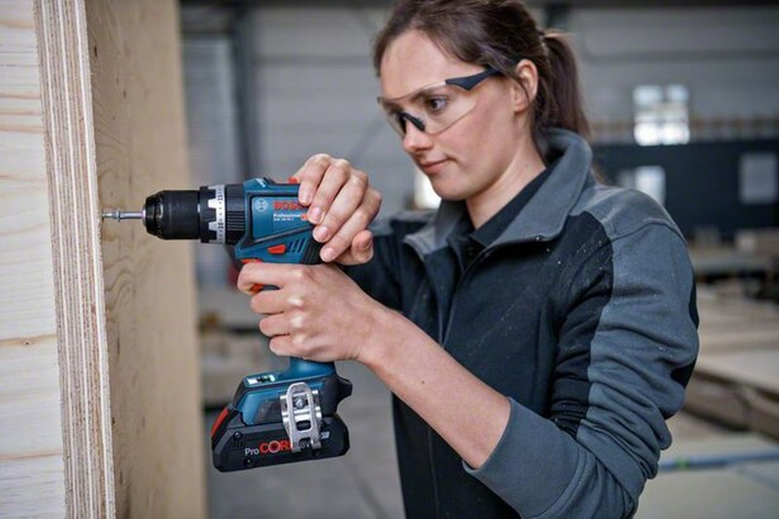 Una donna con occhiali protettivi usa un trapano a batteria blu per avvitare una tavola di legno.