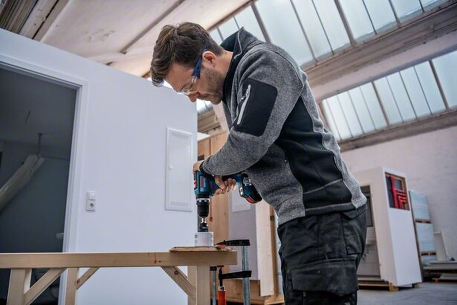 Un artigiano con occhiali protettivi fora il legno in un'officina.