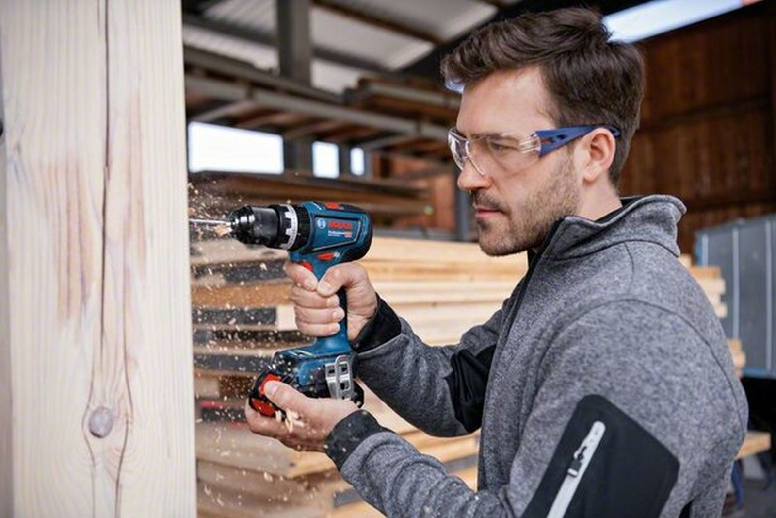 Un artigiano con occhiali protettivi fora una trave di legno mentre vola la segatura.