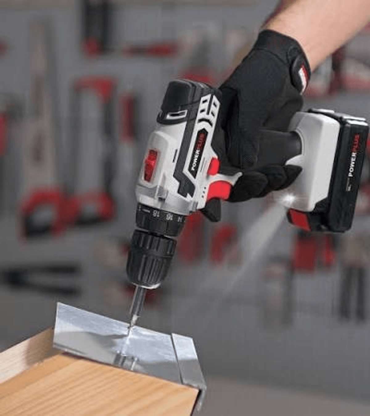 Una mano con guante sostiene un taladro inalámbrico blanco y negro perforando una pieza metálica en madera.