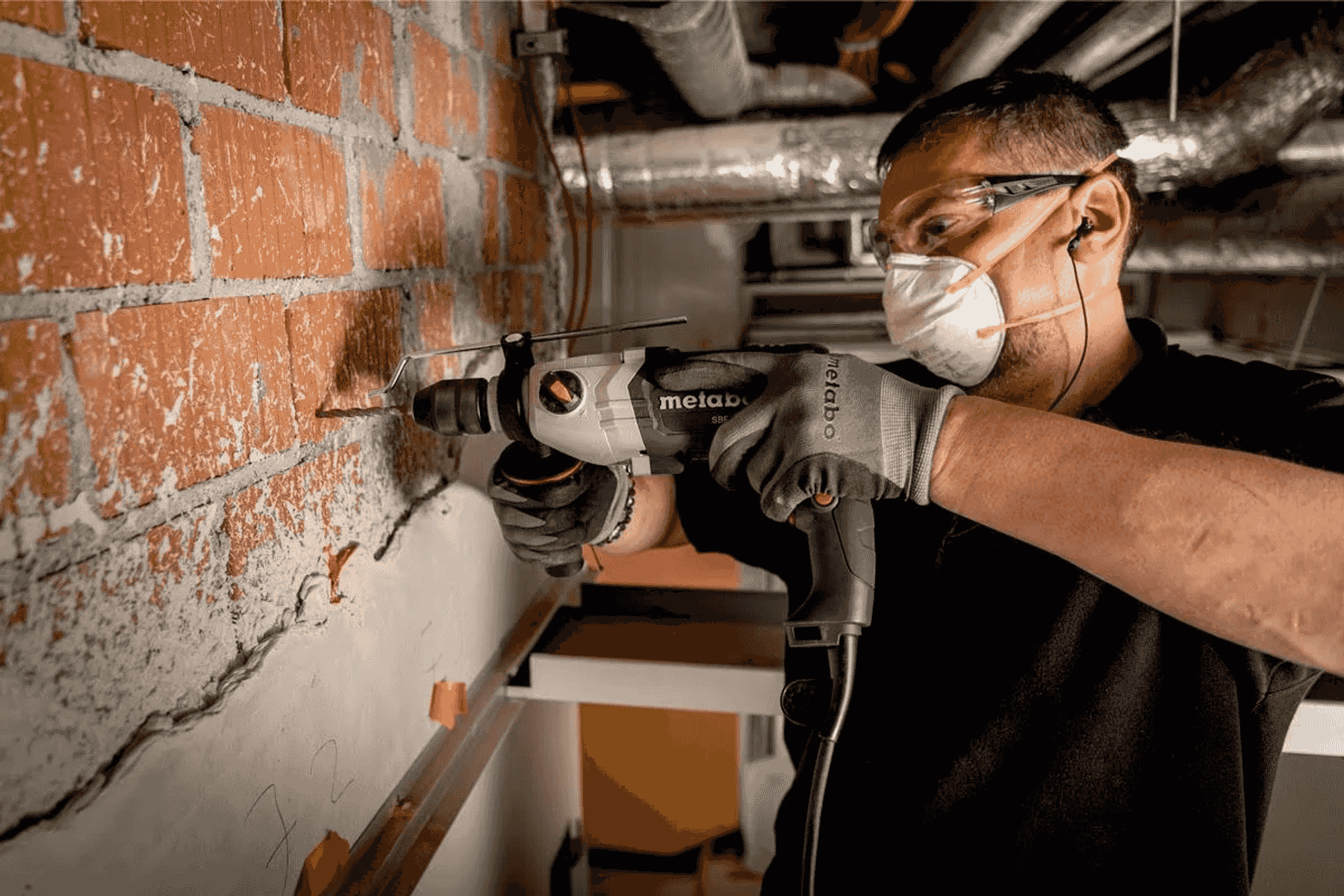 Un trabajador con gafas de seguridad y mascarilla perfora una pared de ladrillo con un taladro Metabo.