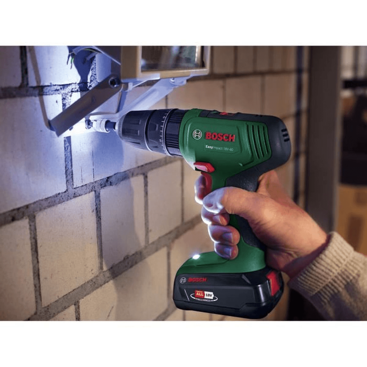 Eine Hand hält einen grünen Bosch Akkubohrer und schraubt eine Leuchte an eine weiße Ziegelwand.