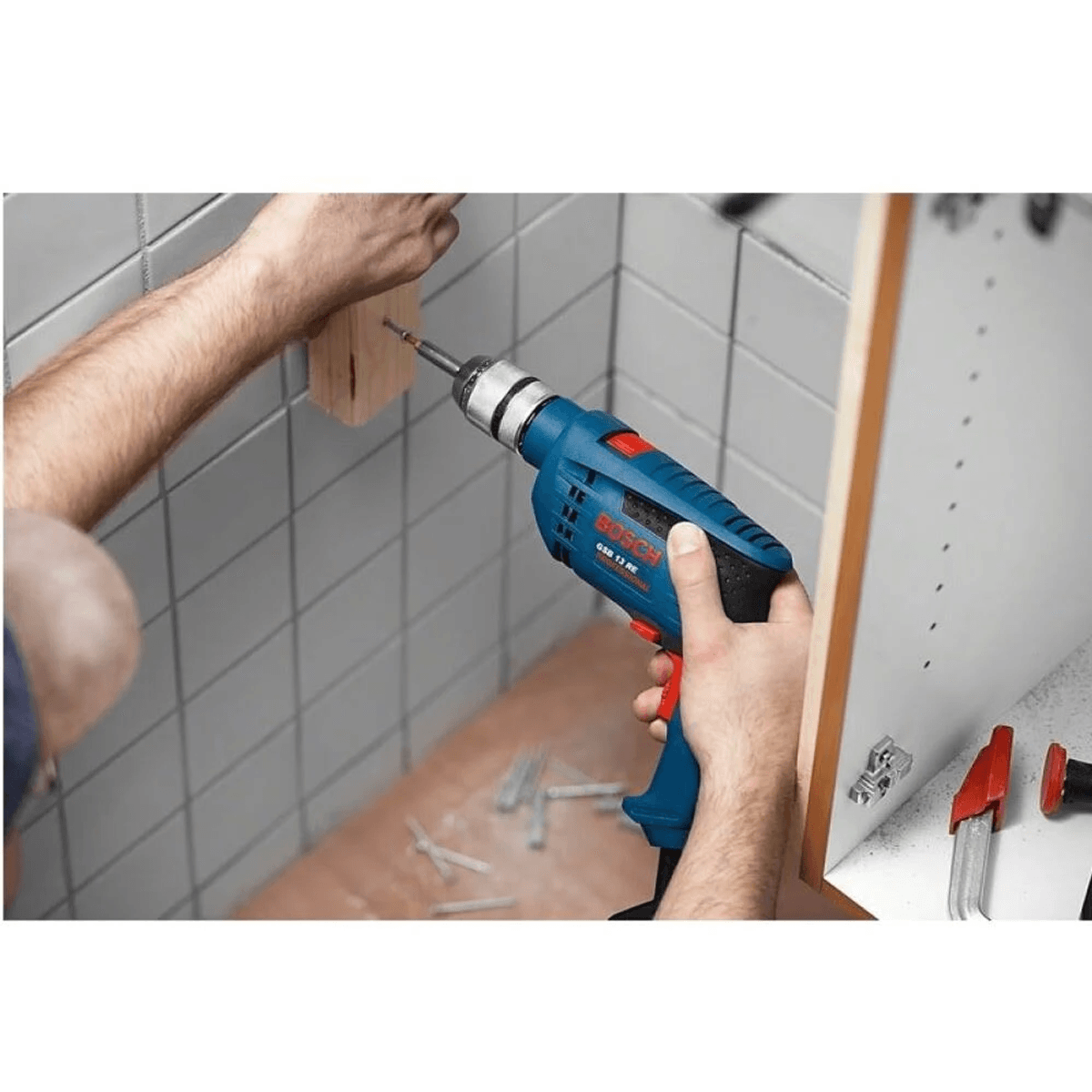 Una persona usa un taladro azul y rojo de Bosch para atornillar un bloque de madera contra una pared de azulejos blancos.