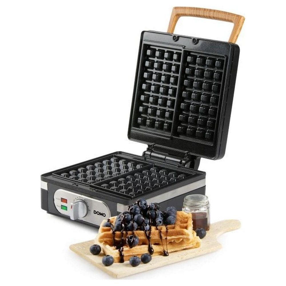 Ein offenes schwarzes Waffeleisen mit Holzgriff steht hinter einem Holzbrett mit Waffeln, Blaubeeren und Sirup.
