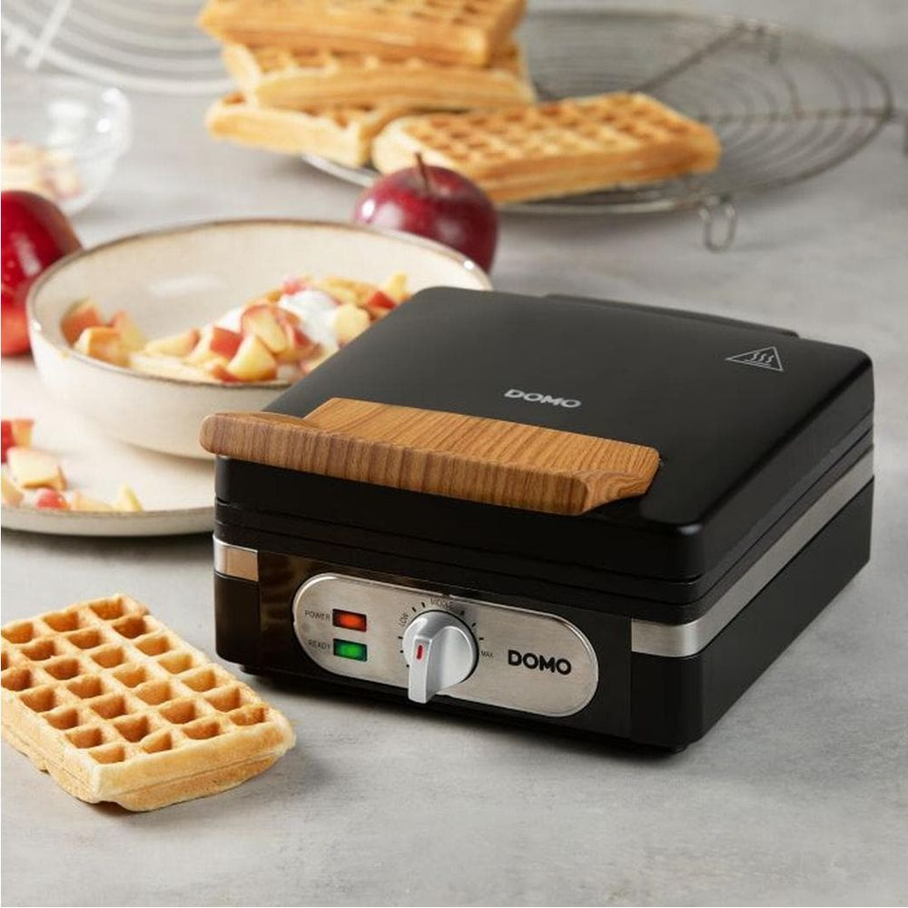 Ein schwarzes Waffeleisen mit Holzgriff steht auf einem Tisch neben Waffeln und Früchten.
