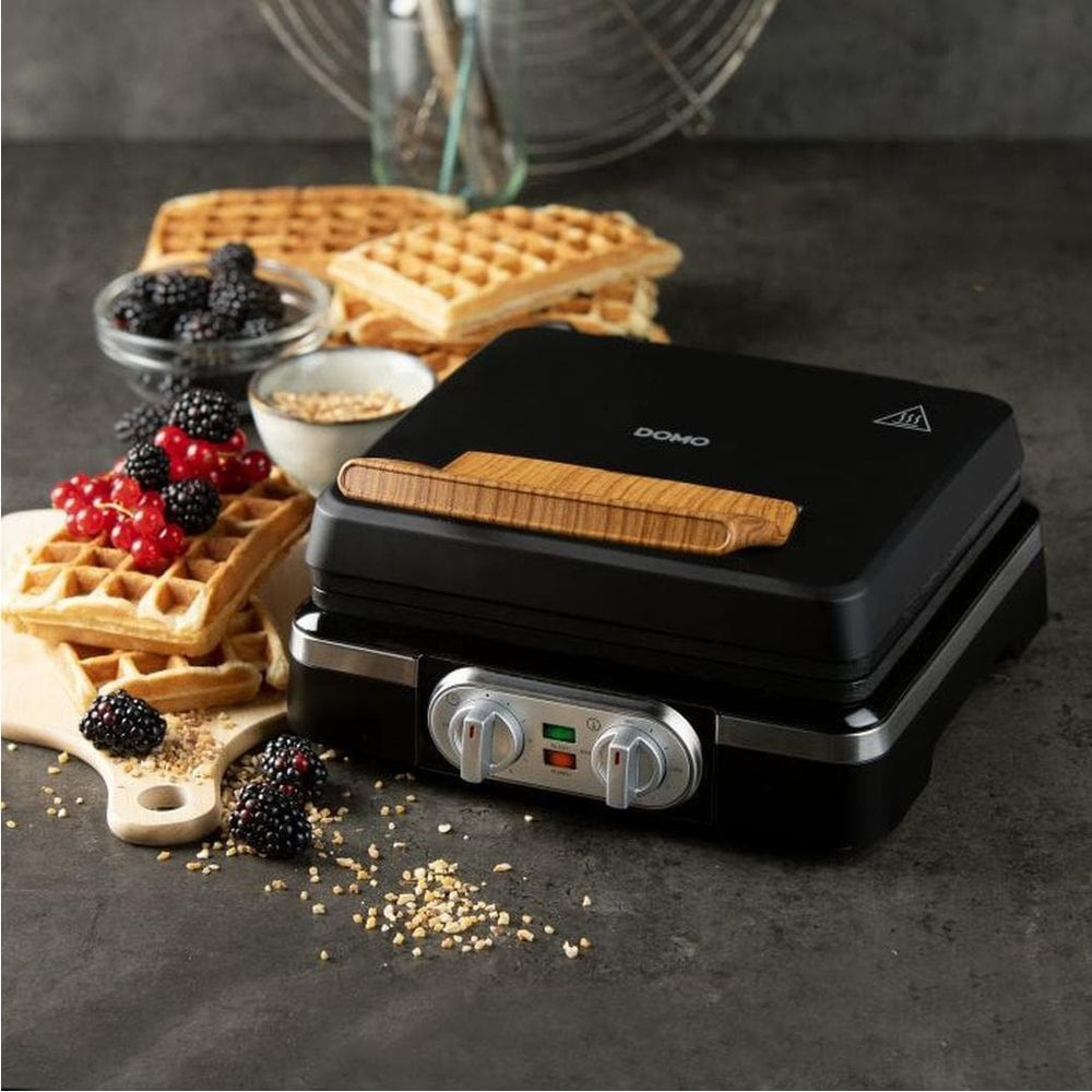 Ein schwarzes Waffeleisen mit Holzgriff steht auf dunklem Tisch, umgeben von Waffeln, Beeren und Körnern.