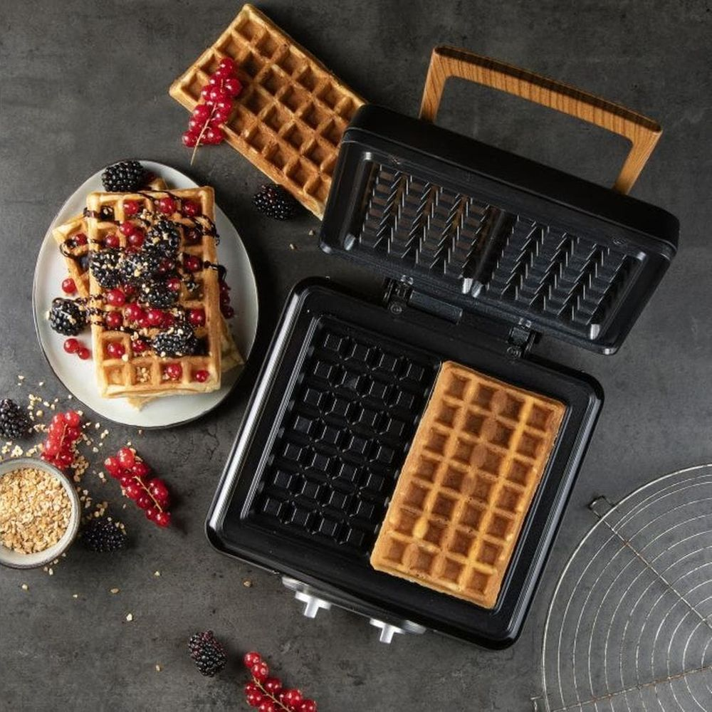 Waffeleisen auf grauem Tisch mit frischen Waffeln, Beeren und Getreide.