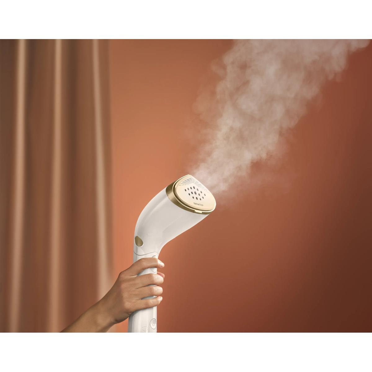 Una mano tiene un vaporizzatore per indumenti bianco e oro, rilasciando vapore su sfondo di tenda marrone.
