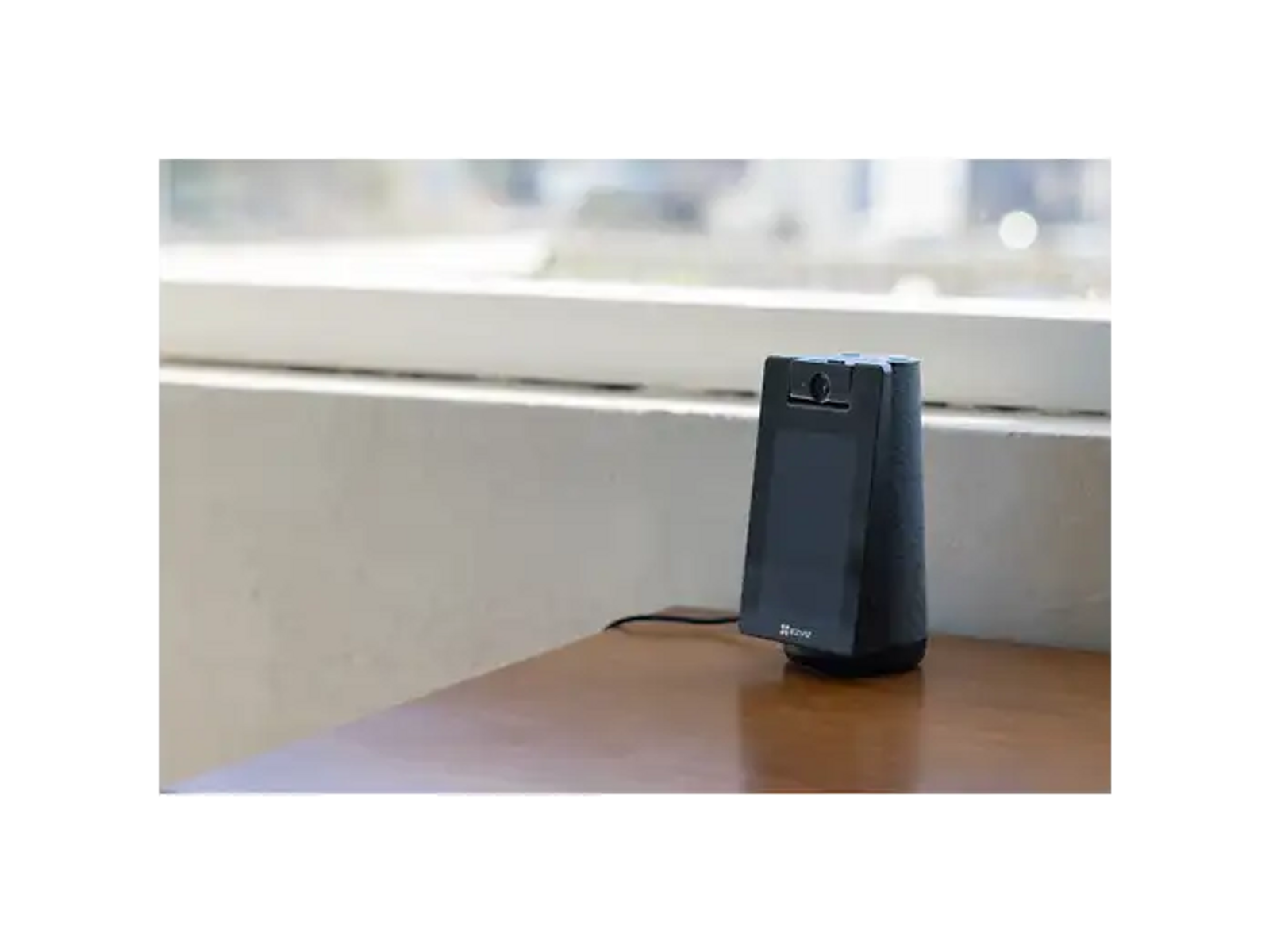A black security device with a camera rests on a wooden table in front of a window.