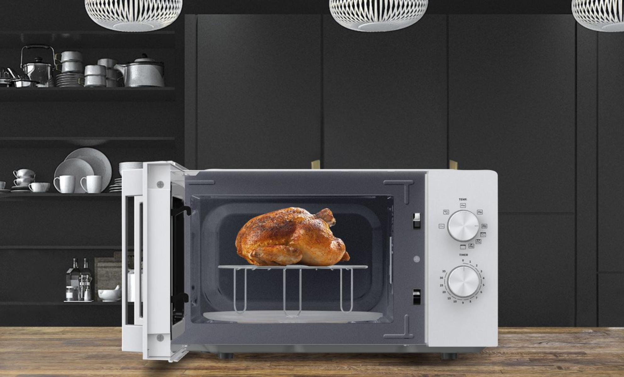 Un forno a microonde bianco su un tavolo di legno con un pollo intero arrosto all'interno, davanti a una cucina scura.