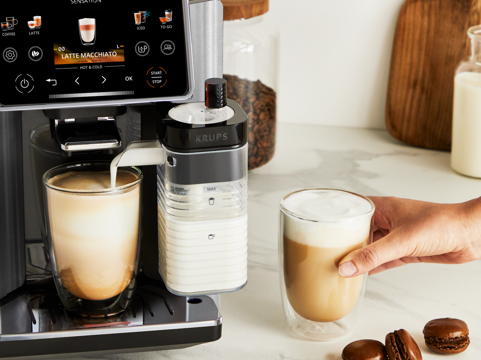 La macchina da caffè Krups versa latte macchiato in un bicchiere, mentre una mano prende un'altra tazza.