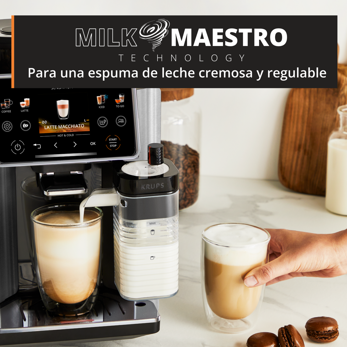 Macchina da caffè Krups versa latte, mano tiene macchiato, chicchi di caffè e macaron sullo sfondo.