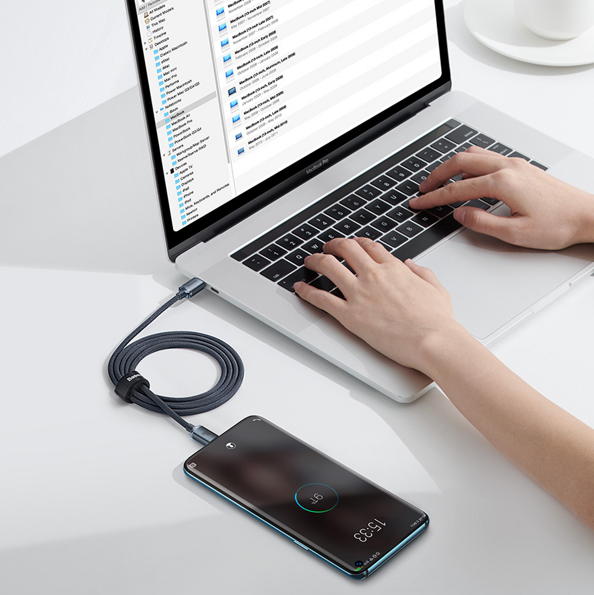 Persoon typt op MacBook Pro, smartphone verbonden via USB-kabel en aan het opladen.