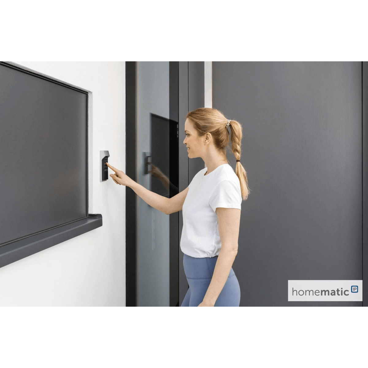 Una mujer con camisa blanca y leggings azules toca un teclado en la pared al lado de una puerta oscura.