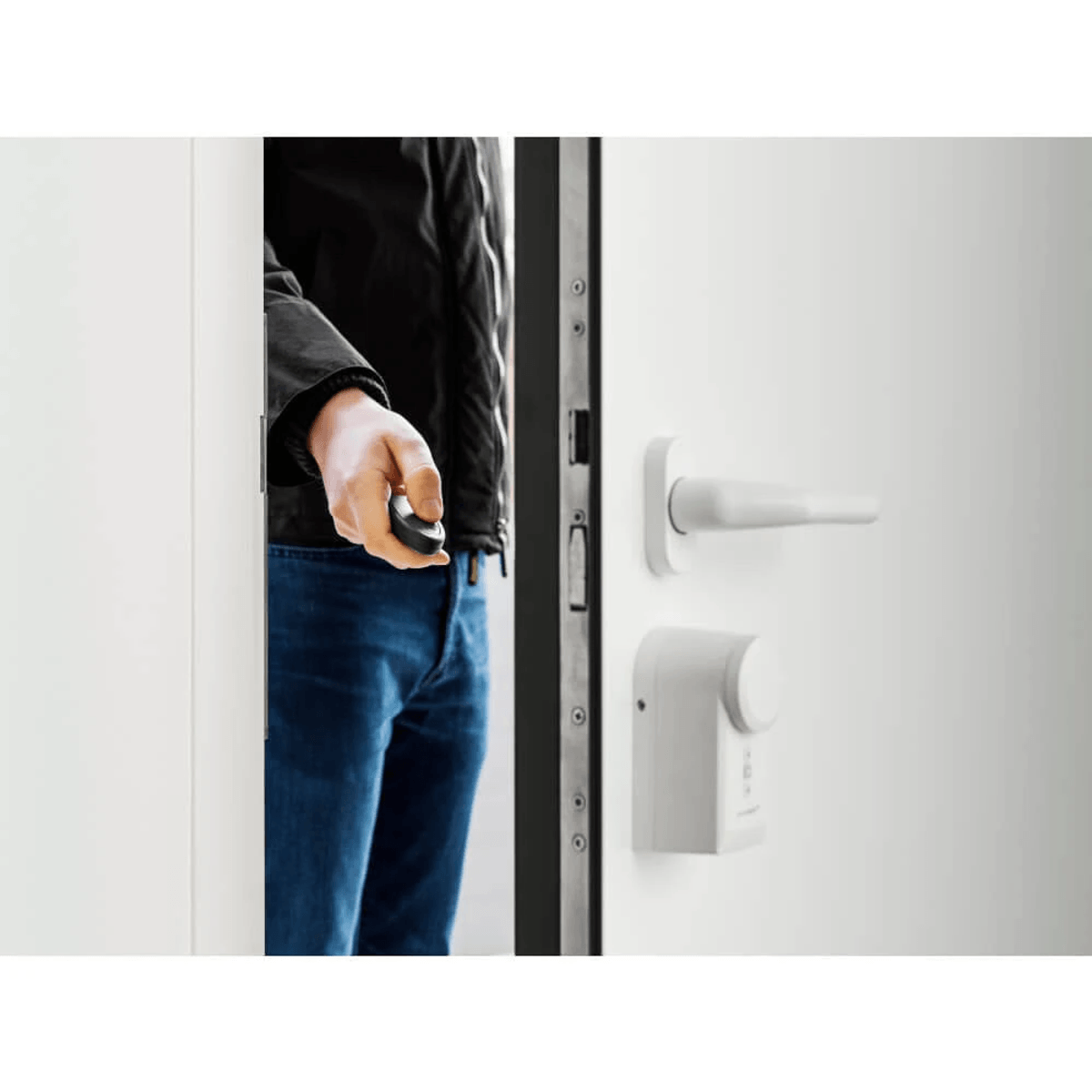 Eine Person in blauen Jeans und schwarzer Jacke öffnet eine weiße Tür mit elektronischem Schloss und Fernbedienung.
