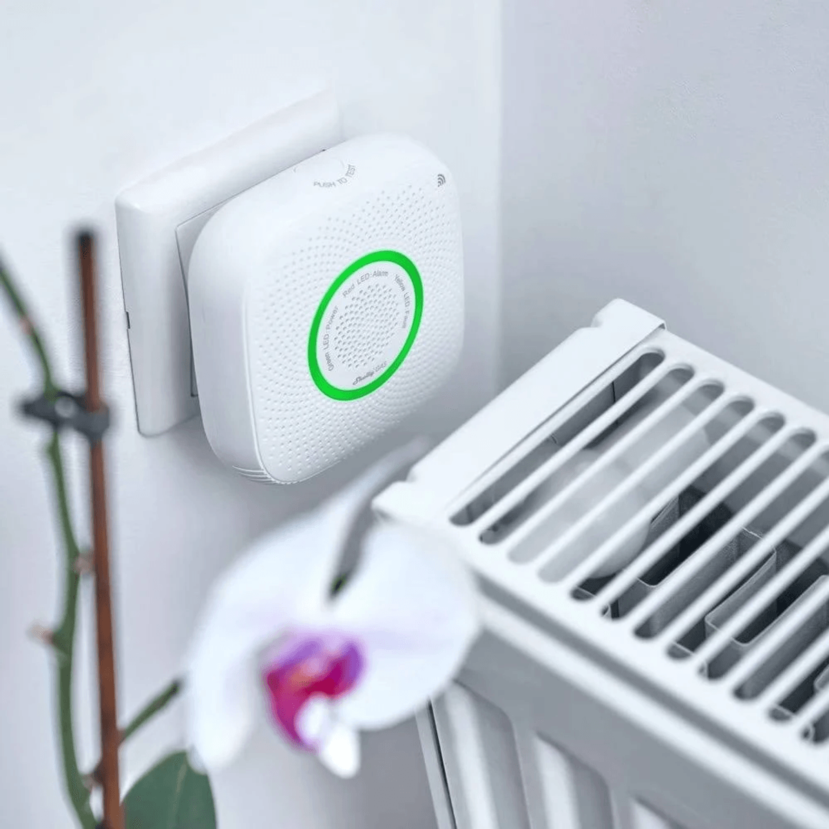 Un détecteur de gaz blanc avec un anneau LED vert est branché sur une prise murale près d'un radiateur.