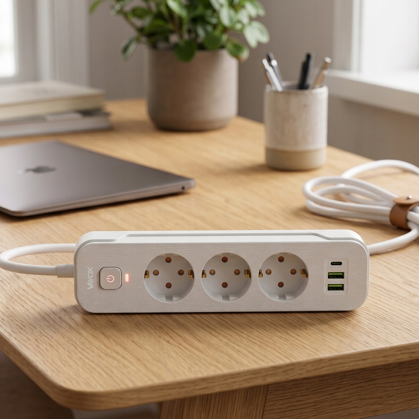 Biała listwa zasilająca z trzema gniazdami i portami USB leży na drewnianym biurku, w tle laptop.