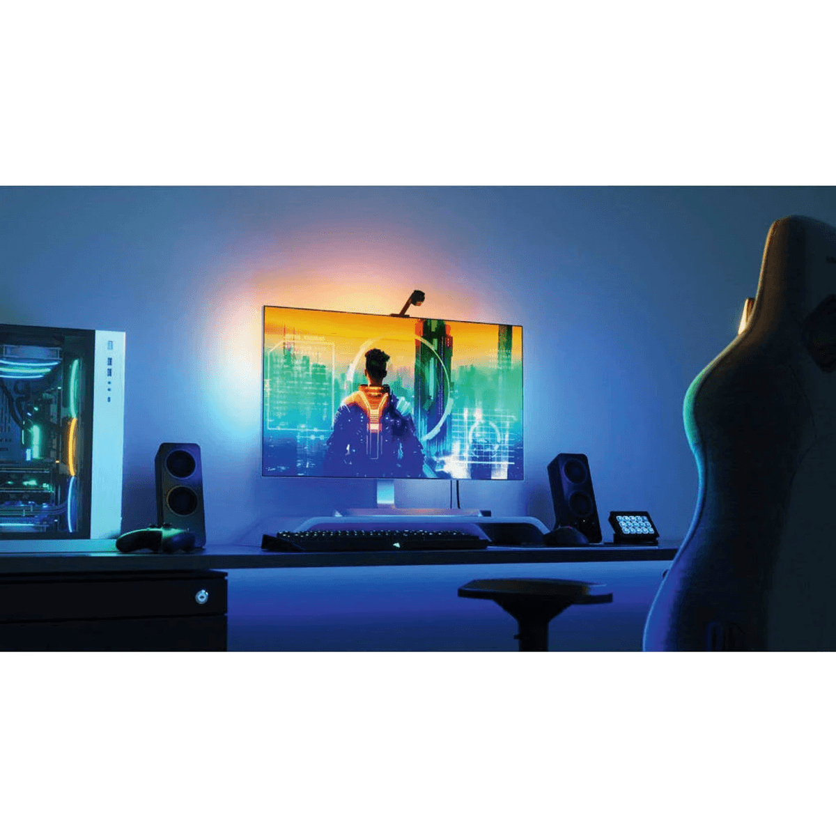 Configuración de juegos con monitor iluminado, PC, altavoces, teclado y silla frente a una pared azul.