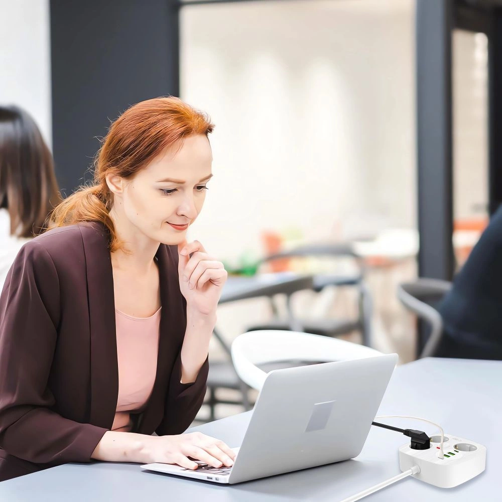 Eine Frau arbeitet an einem Laptop an einem Tisch, im Vordergrund ist eine weiße Steckerleiste angeschlossen.