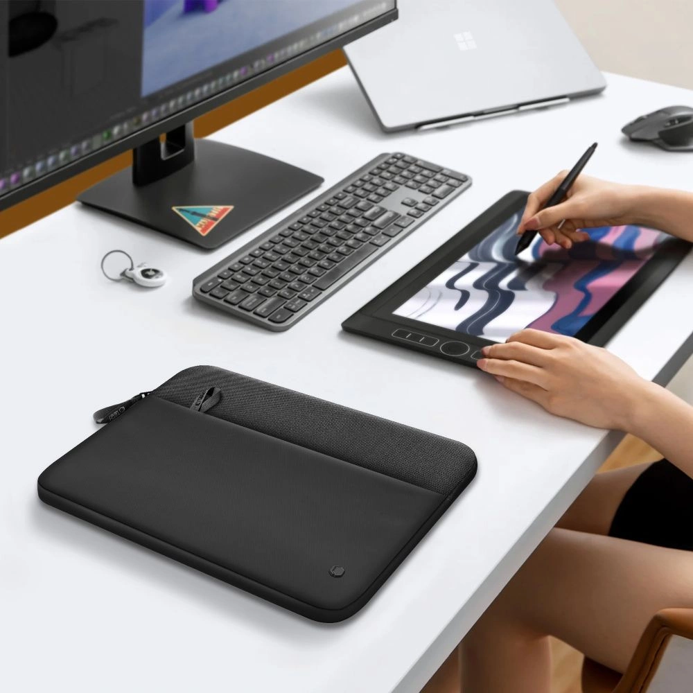 A person draws on a graphics tablet at a white desk with a monitor and a laptop sleeve.