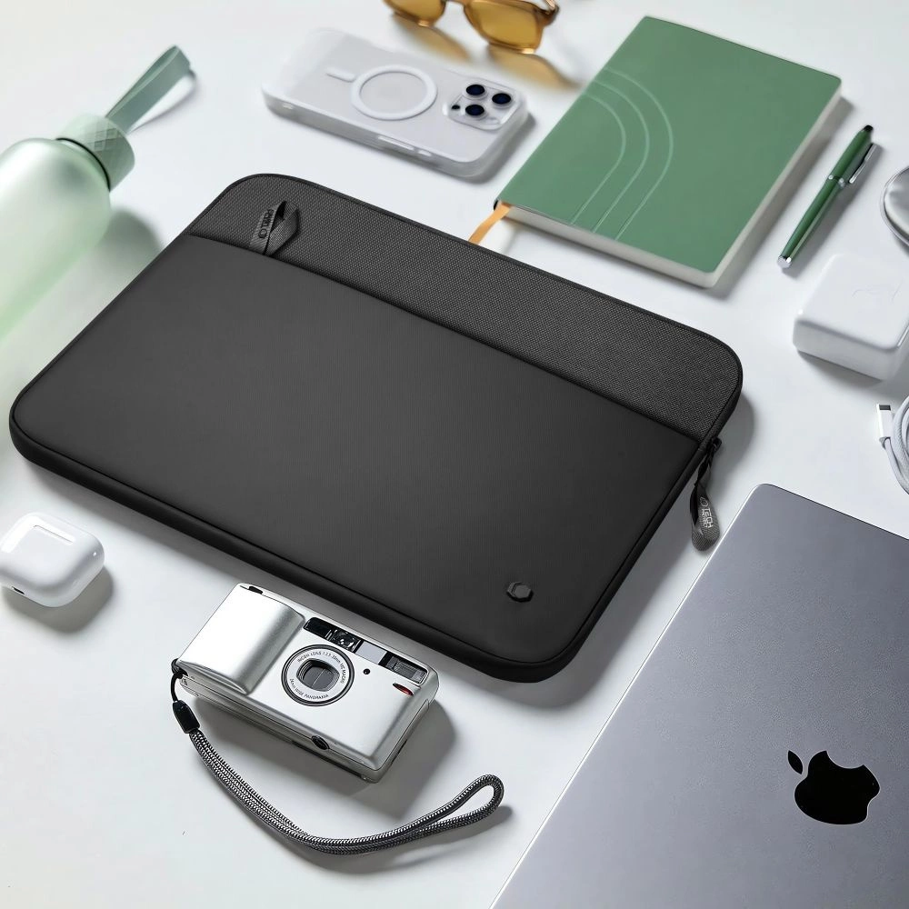 A black laptop sleeve on a white surface, surrounded by a camera, phone, green notebook, pen, and water bottle.