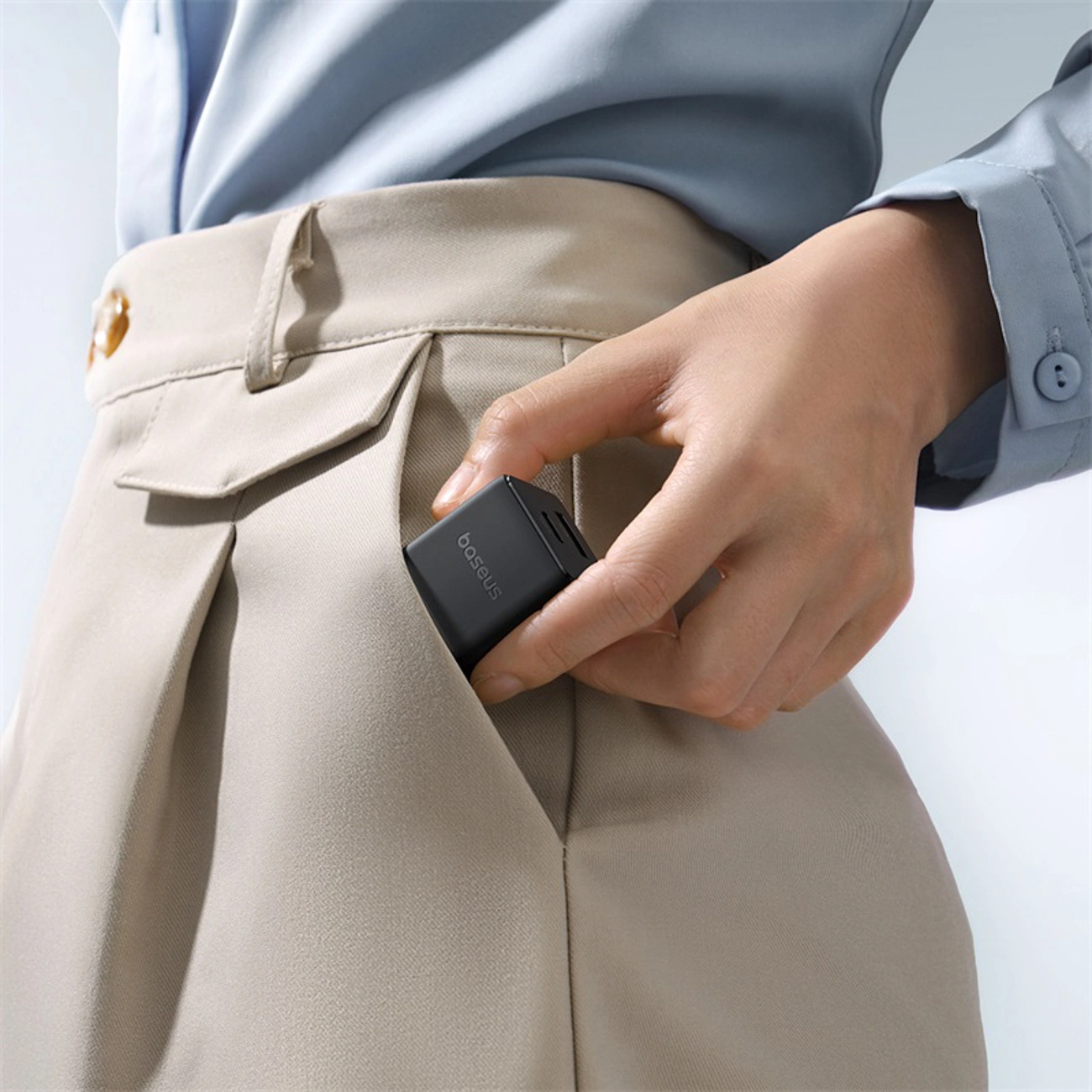 Une personne en chemise bleu clair et pantalon beige range un petit chargeur noir Baseus dans sa poche.