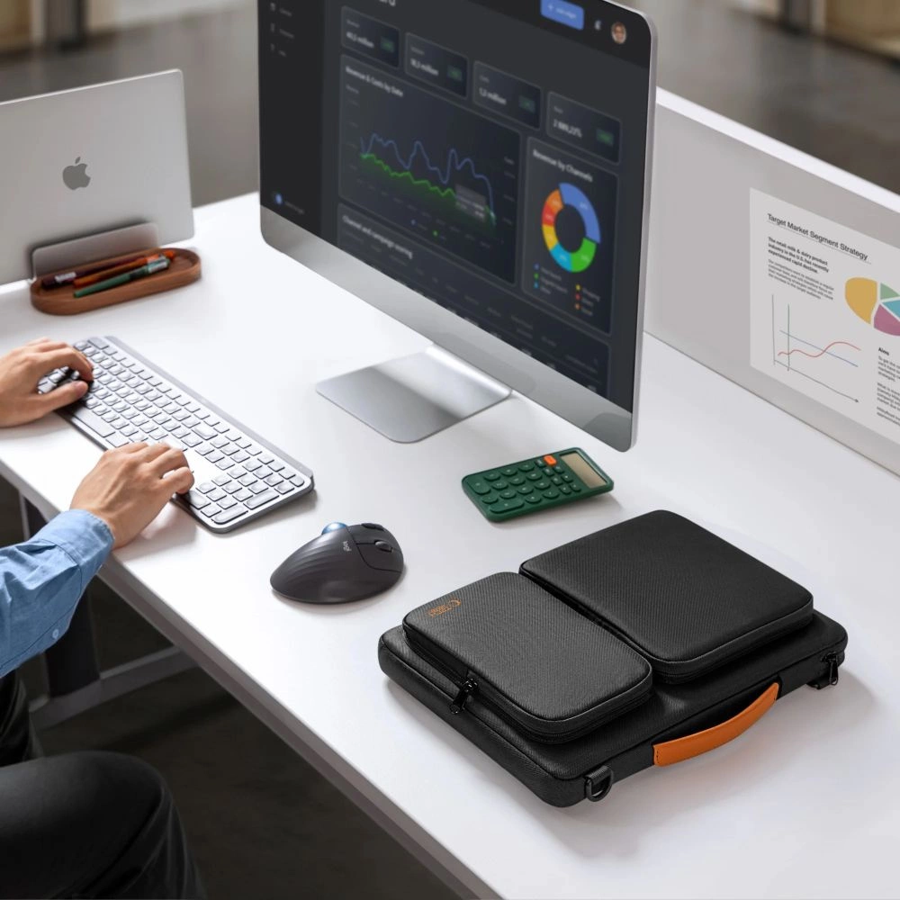 Person tippt auf weißem Schreibtisch mit Monitor, Laptop, Taschenrechner, Maus und schwarzer Laptoptasche.