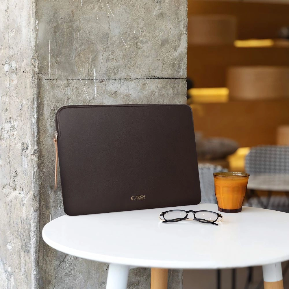 Een bruine laptophoes, een bril en een glas jus d'orange op een witte tafel tegen een grijze betonnen muur.