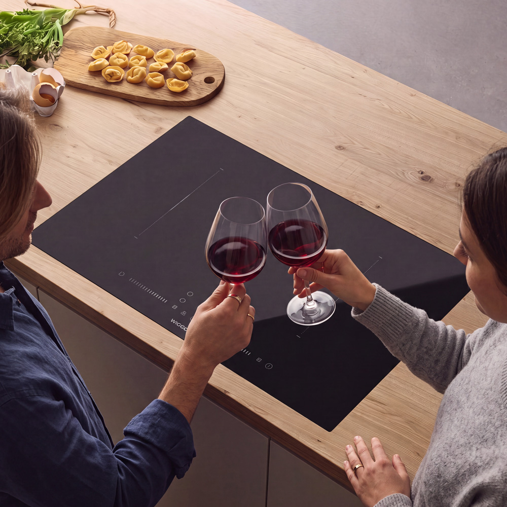 Een koppel proost met rode wijnglazen boven een kookplaat, met ravioli en groenten op een houten blad.