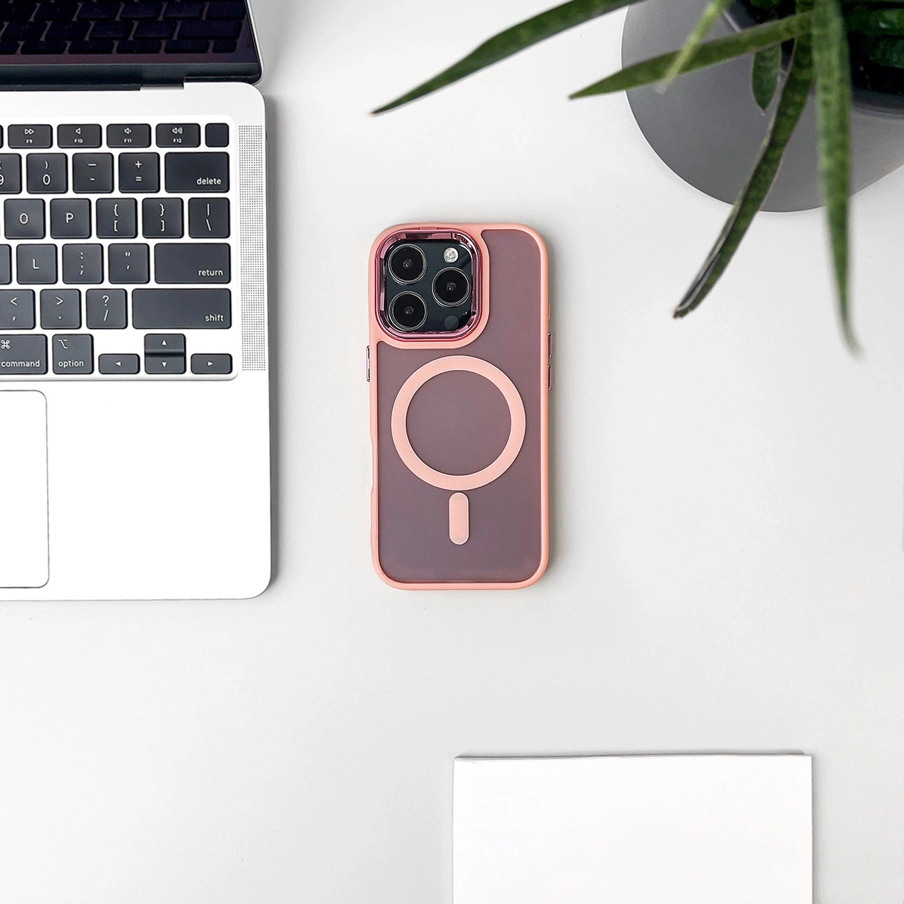 Eine rosa MagSafe-Handyhülle auf einem weißen Schreibtisch neben einem Laptop, Topfpflanze und Notizbuch.