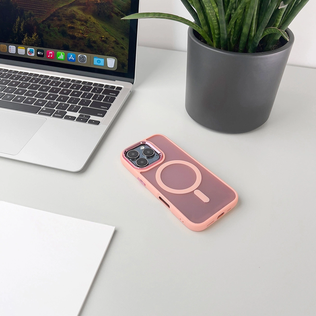 Eine rosa iPhone-Hülle, ein Laptop, ein Papier und eine Pflanze im Topf auf einem Tisch.