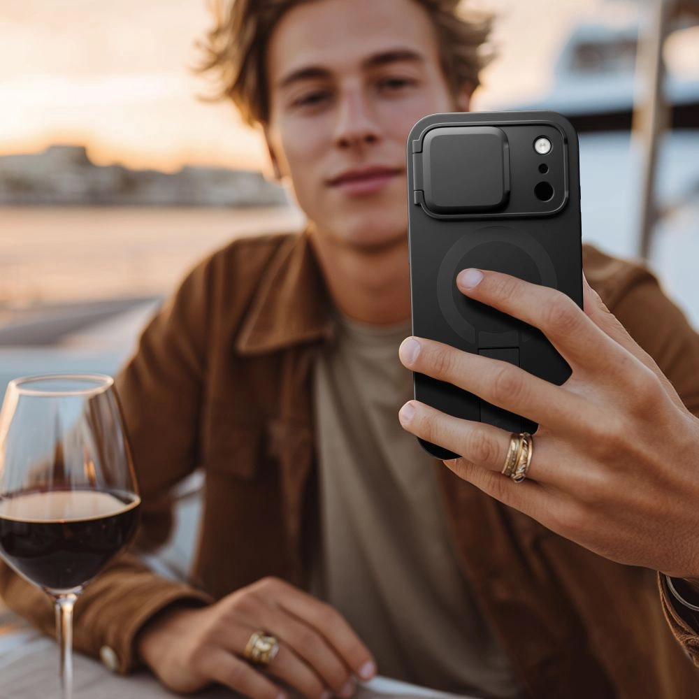 Ein junger Mann im braunen Hemd hält ein schwarzes Smartphone mit Kameraabdeckung vor sich, links steht ein Weinglas.