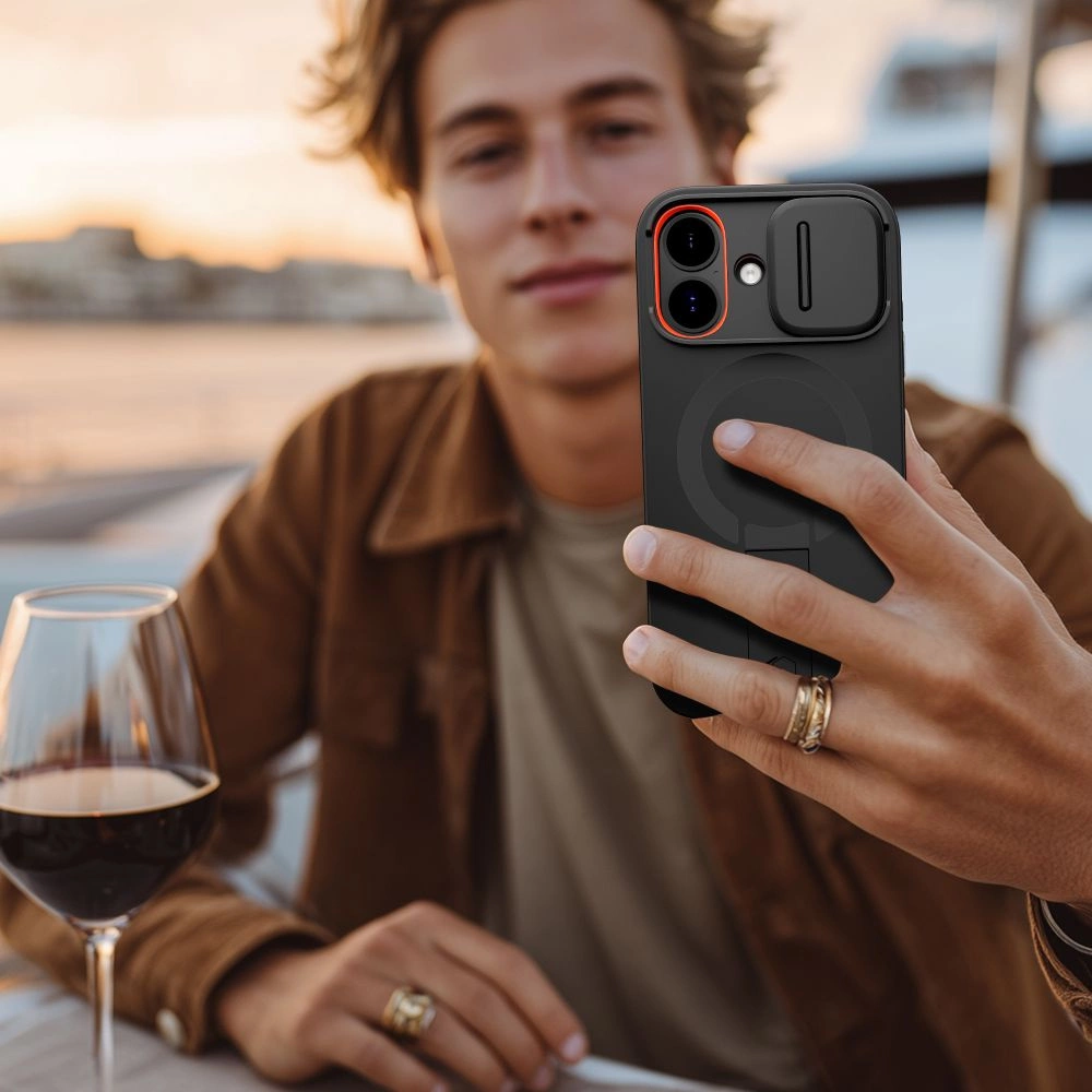 Ein junger Mann hält ein Smartphone mit schwarzer Hülle, im Hintergrund sind ein Weinglas und ein See zu sehen.