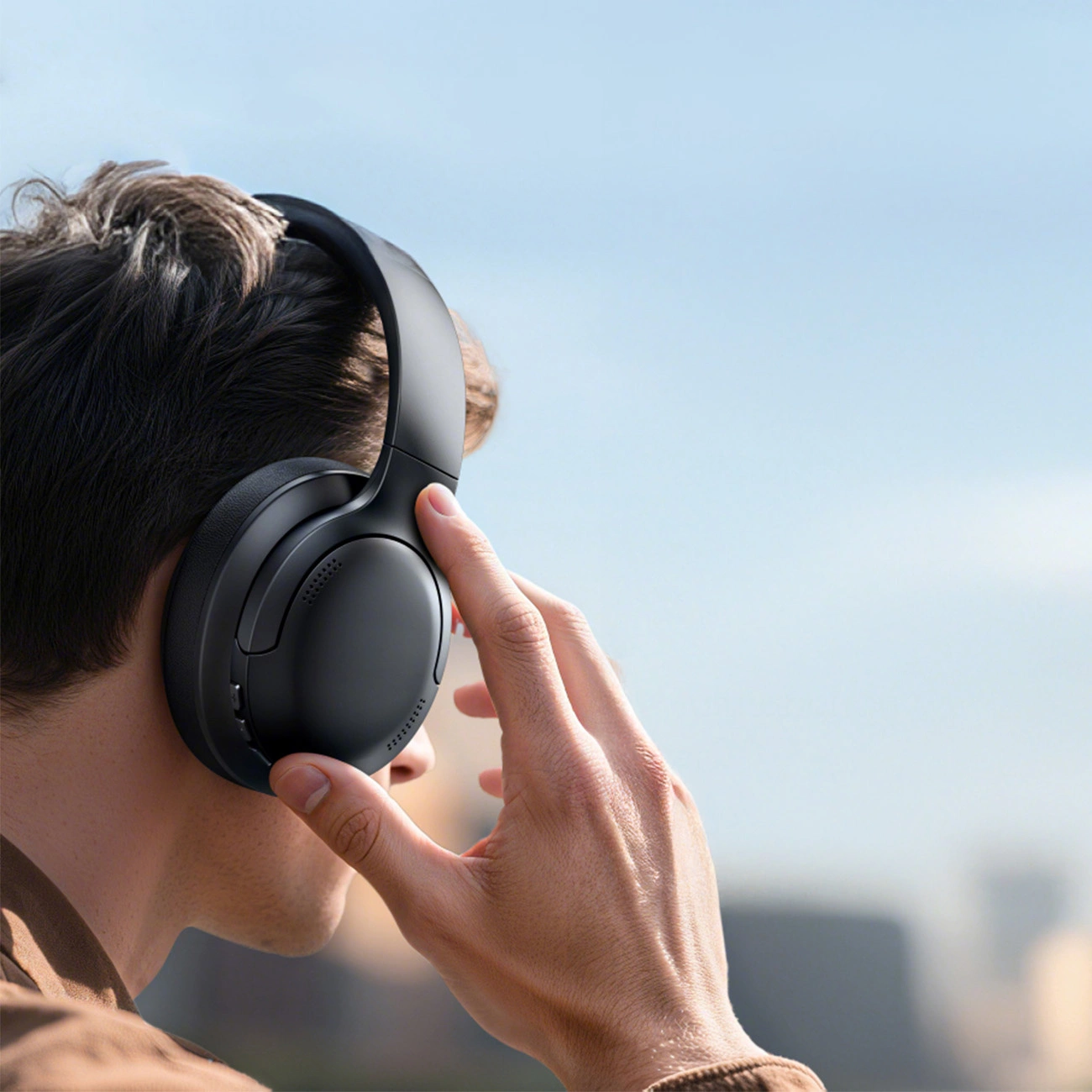 Un uomo con i capelli castani indossa cuffie nere over-ear, toccando il padiglione auricolare su sfondo chiaro.
