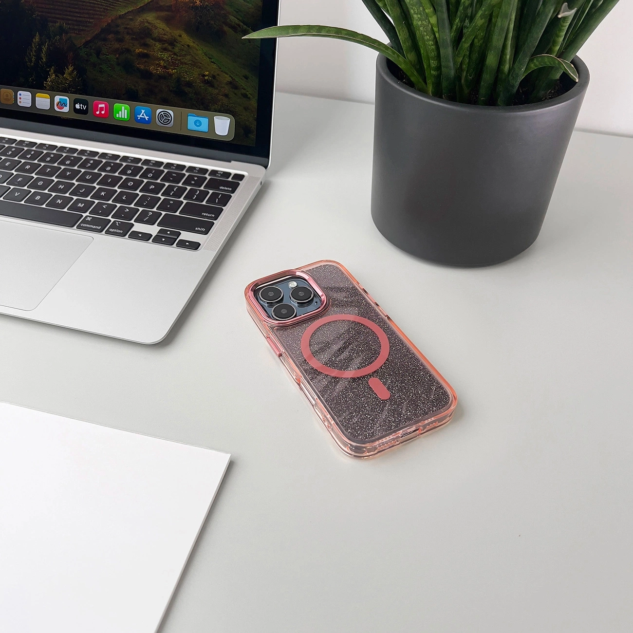Ein glitzerndes rosafarbenes iPhone in Hülle auf weißem Schreibtisch neben Laptop und Pflanze.