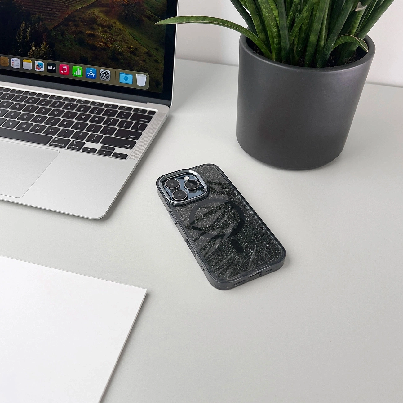 Ein Smartphone mit glitzernder Schutzhülle liegt neben einem Laptop, einer Pflanze und Papier auf einem Tisch.
