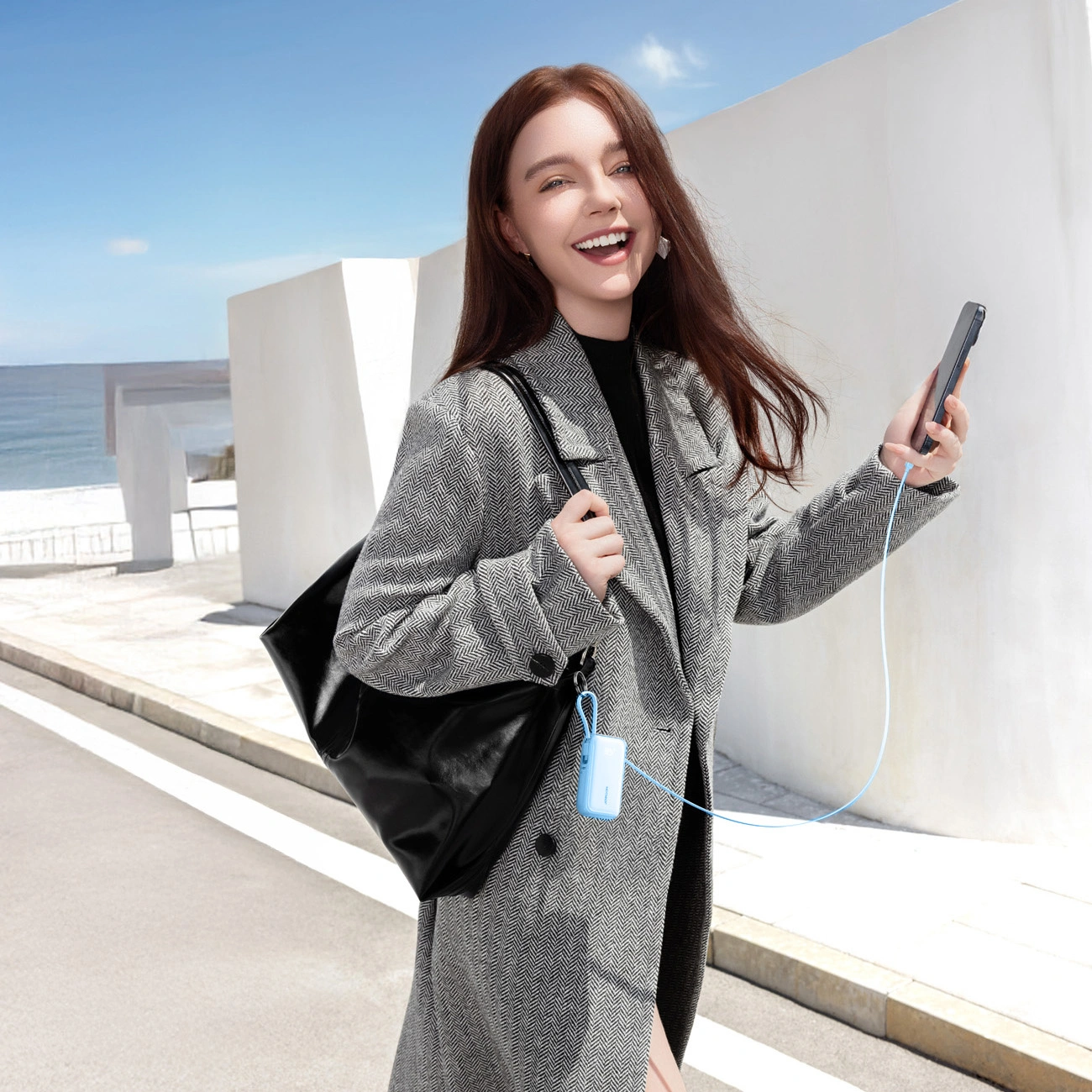 Donna sorridente in cappotto grigio con borsa nera che tiene uno smartphone collegato con un cavo blu.