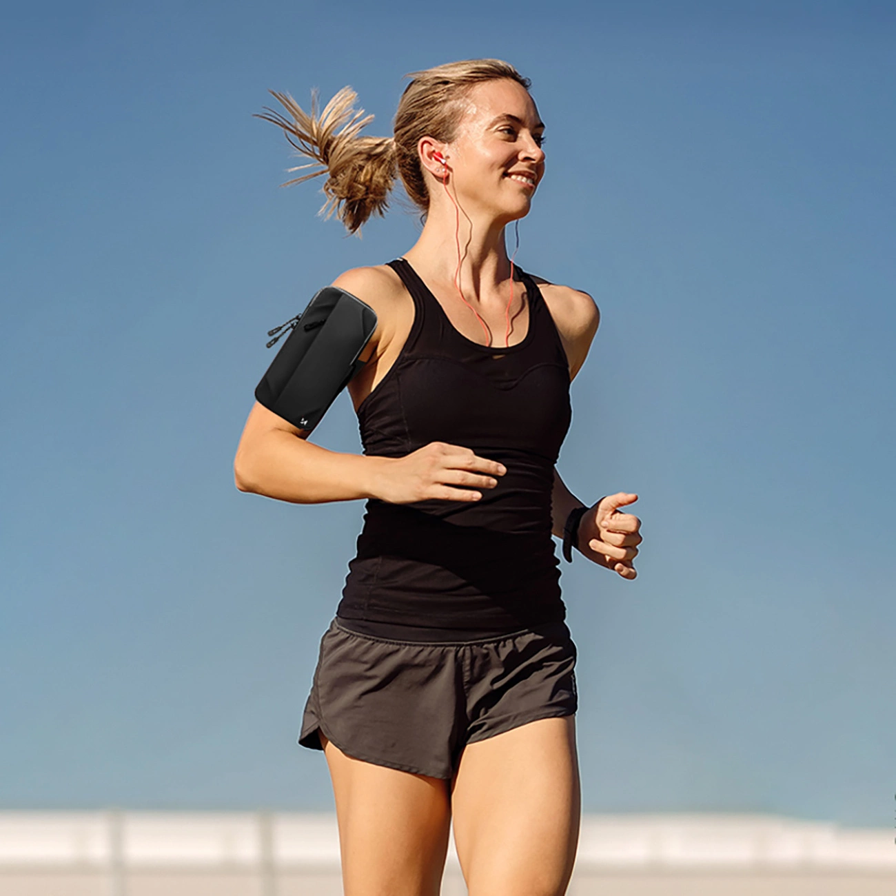 Eine blonde Frau in Sportkleidung läuft im Freien vor blauen Himmel, trägt ein schwarzes Armband und rote Kopfhörer.