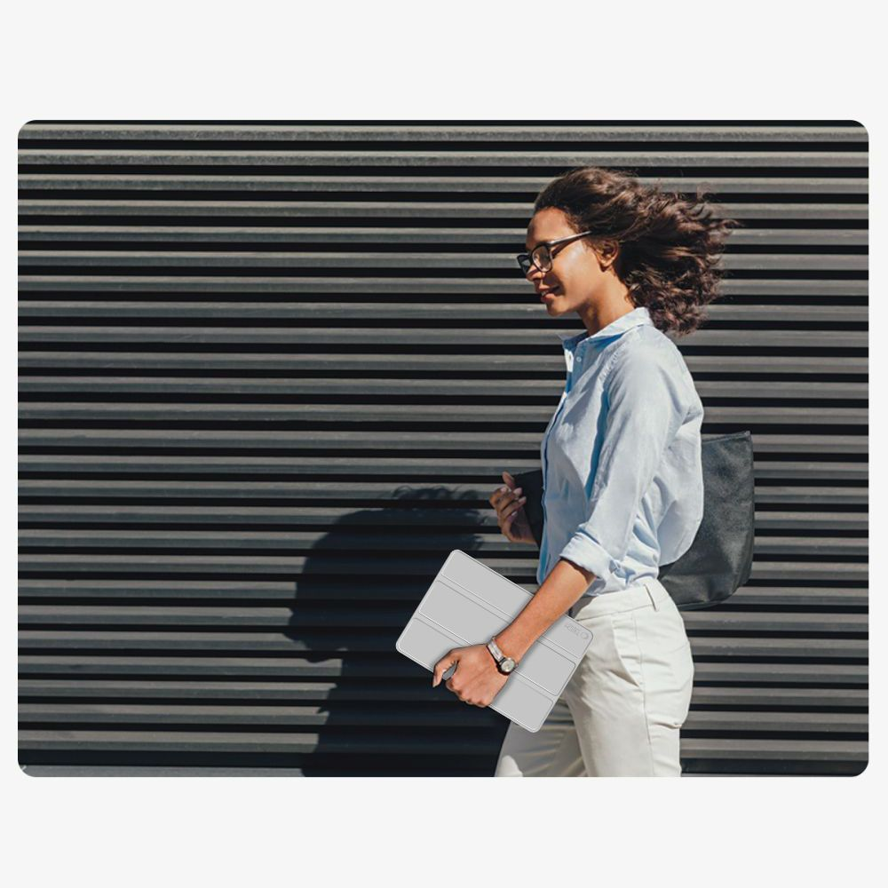 Una donna con camicia blu e pantaloni chiari cammina con un tablet davanti a una parete scura.