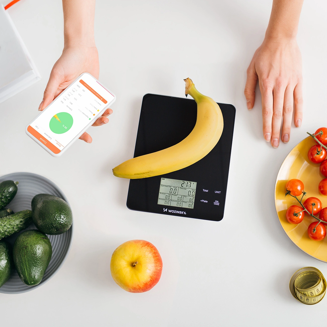 Una banana riposa su una bilancia da cucina digitale, circondata da frutta, un'app e un metro.