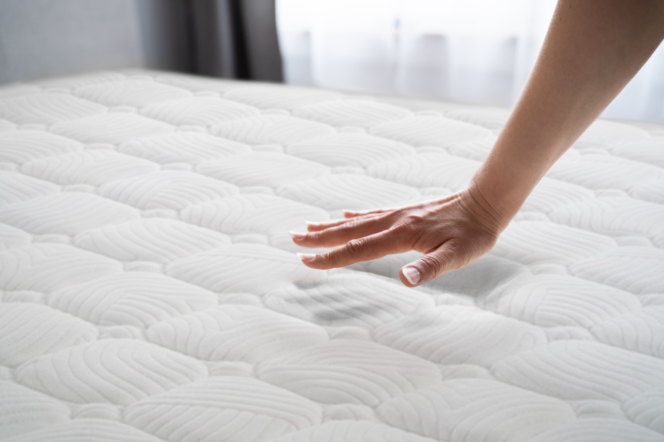 Una mano presiona un colchón blanco y acolchado en una habitación luminosa.