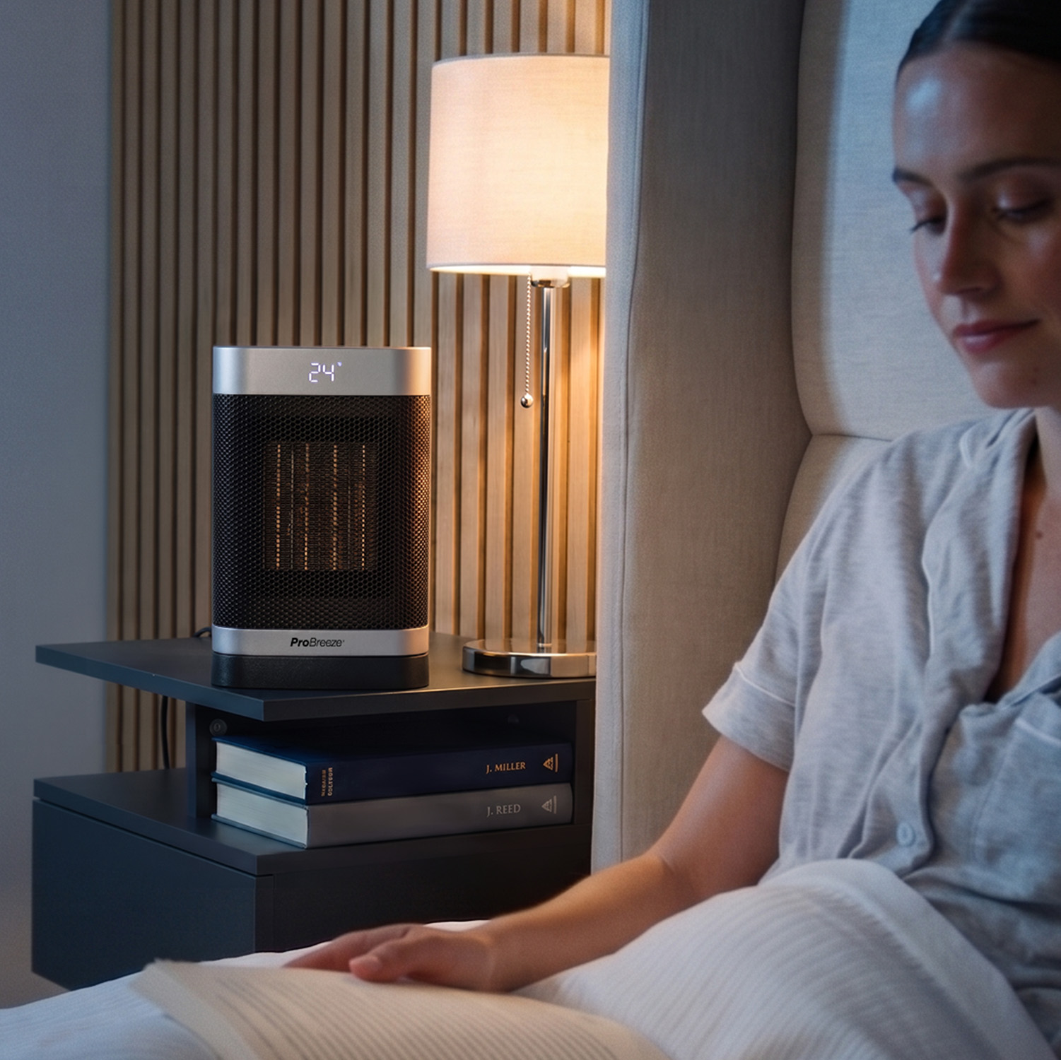 Eine Frau liest im Bett, neben ihr auf dem Nachttisch steht ein Heizlüfter.