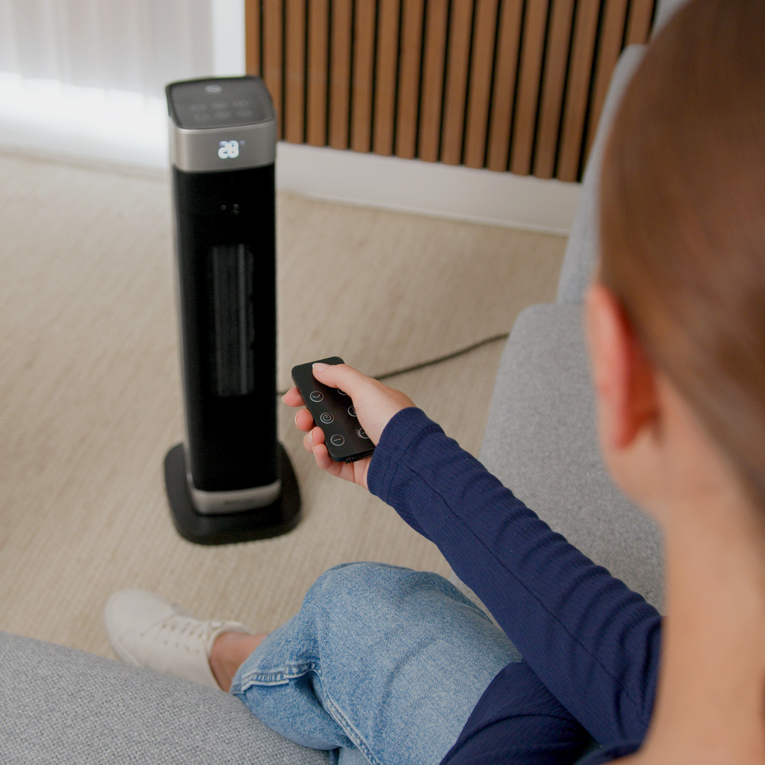 Eine Frau sitzt auf einem Sofa und steuert eine schwarze Heizsäule mit einer Fernbedienung.