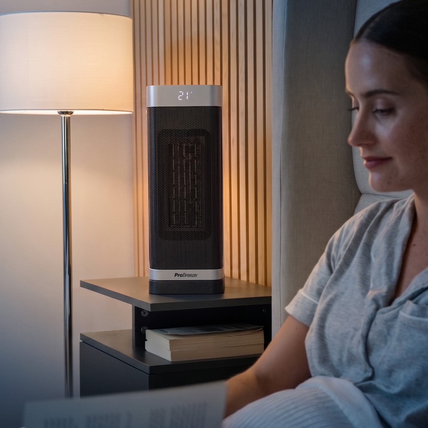 Eine Frau liest im Bett, neben ihr auf einem Nachttisch steht ein Turmheizlüfter, im Hintergrund eine Lampe.
