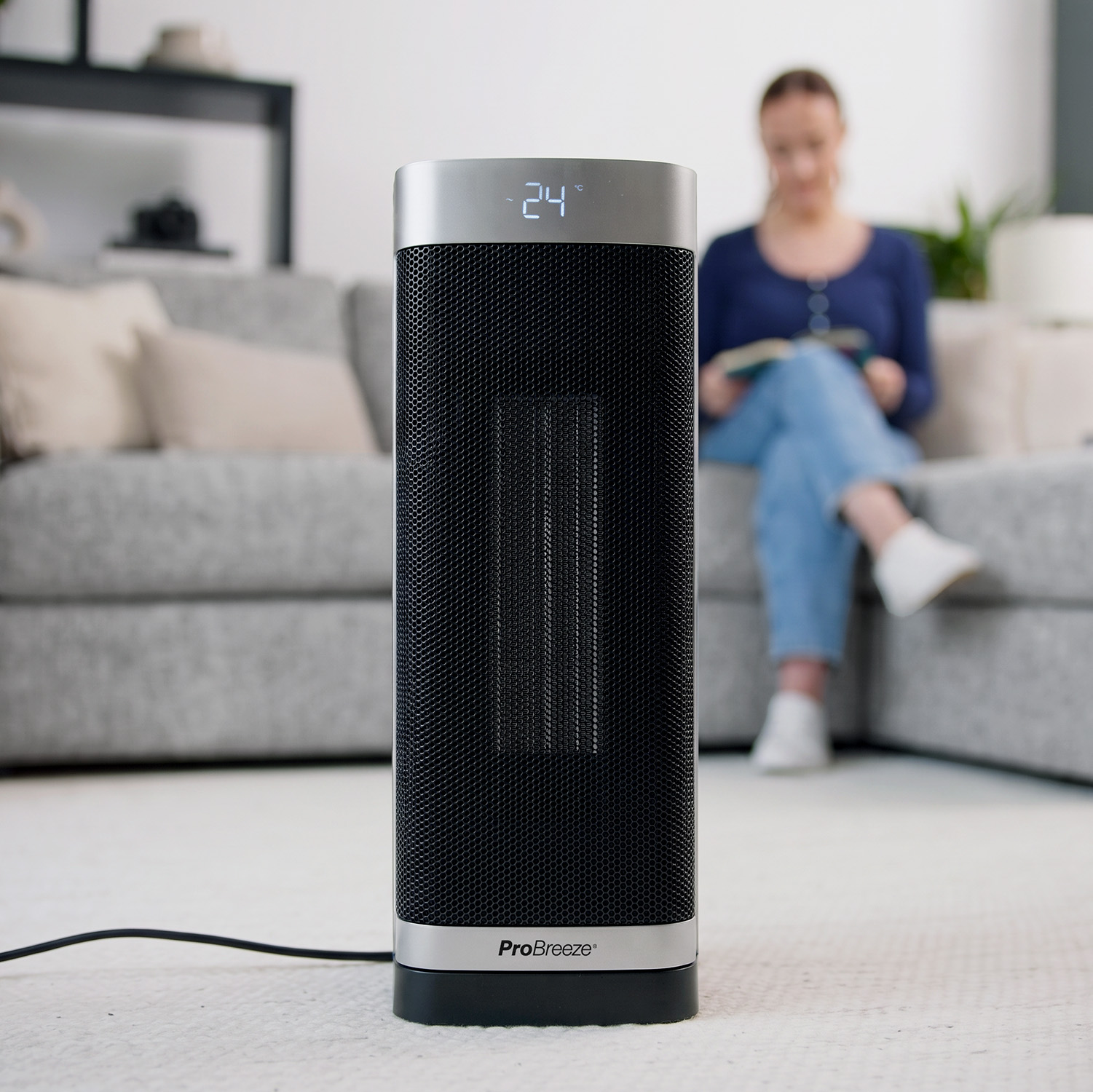 Ein schwarzer ProBreeze Turmventilator mit digitaler Anzeige auf einem Teppich in einem Wohnzimmer, eine Frau sitzt im Hintergrund.