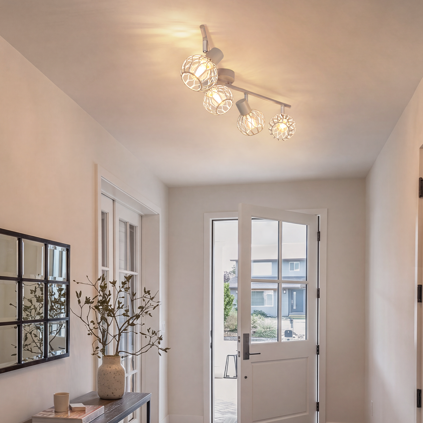 Un pasillo luminoso con foco de techo blanco, puerta principal abierta y mesa con planta.