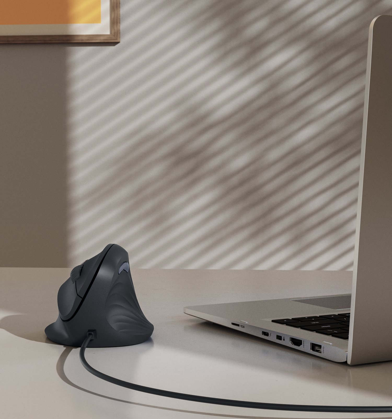 Un mouse verticale ergonomico grigio con cavo accanto a un computer portatile su una scrivania con ombre.