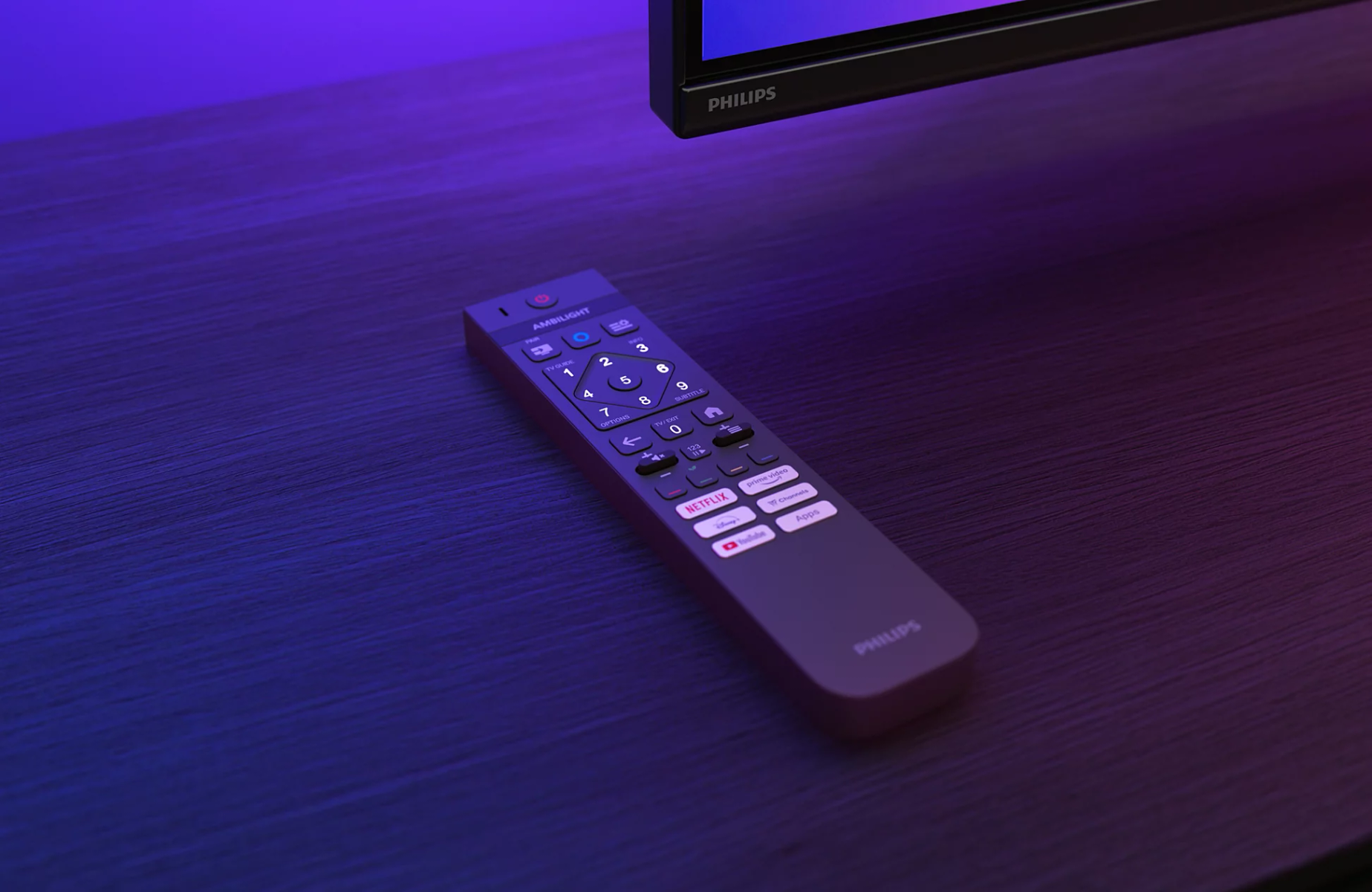 Pilot Philips na ciemnej drewnianej powierzchni pod telewizorem w niebieskim świetle otoczenia.