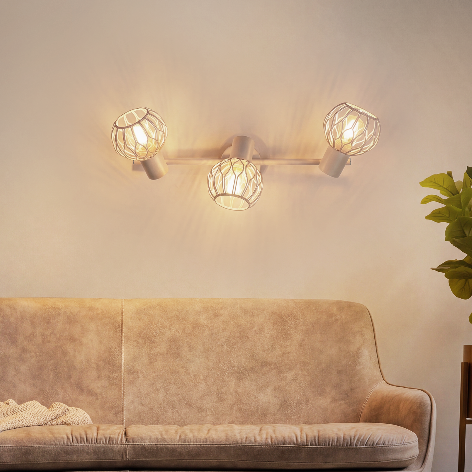 Beigefarbene Couch unter einer modernen dreiflammigen Wandlampe an einer hellen Wand, rechts eine Grünpflanze.