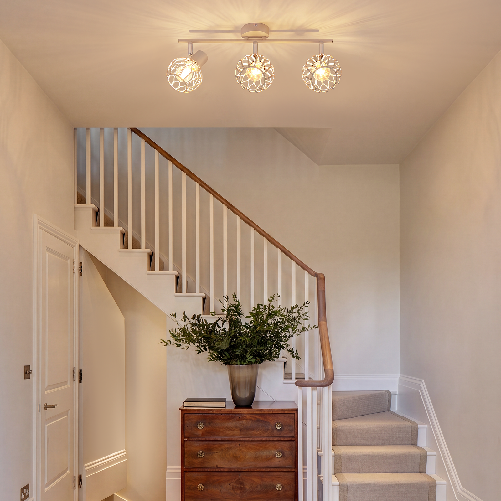 Ein heller Flur mit Holzkommode, grüner Pflanze, Treppe und moderner Deckenleuchte.
